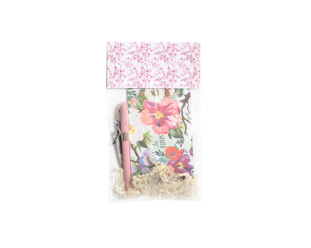 A floral stationery gift set packaged in a clear plastic bag with a decorative pink and white floral top closure. The set includes a vibrant floral art print featuring colorful flowers and the words 'les FLEURS,' a pink fountain pen with silver accents, and crinkle-cut paper filler at the bottom for added presentation.