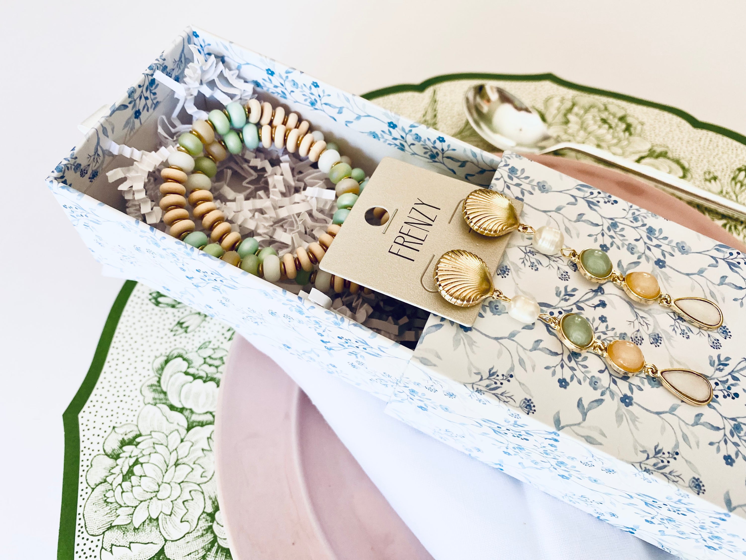 Gift box displaying Frenzy brand gold shell earrings and a green and gold beaded bracelet, presented on a white and green decorative table setting.