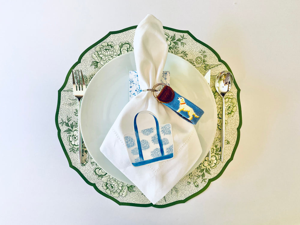 Elegant table setting featuring a green and white floral-patterned plate, white napkin with a blue handbag design, and a keychain with a dog motif, placed on a white background with silver utensils