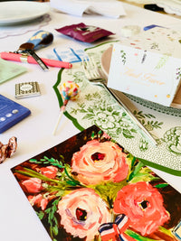 Festive table setting with a green and white floral plate, pink plate, and various items including a floral card, lollipop, and Kind Favor gift box, arranged on a white tablecloth