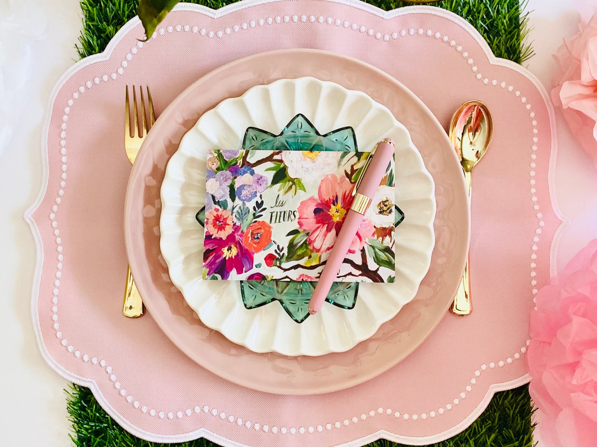 Elegant table setting with a pink scalloped placemat, white plate, and a decorative green glass plate topped with a floral card and a pink pen. The setup is complemented by gold cutlery and a grass mat for added texture and freshness. The arrangement exudes a charming and sophisticated ambiance perfect for a special occasion.