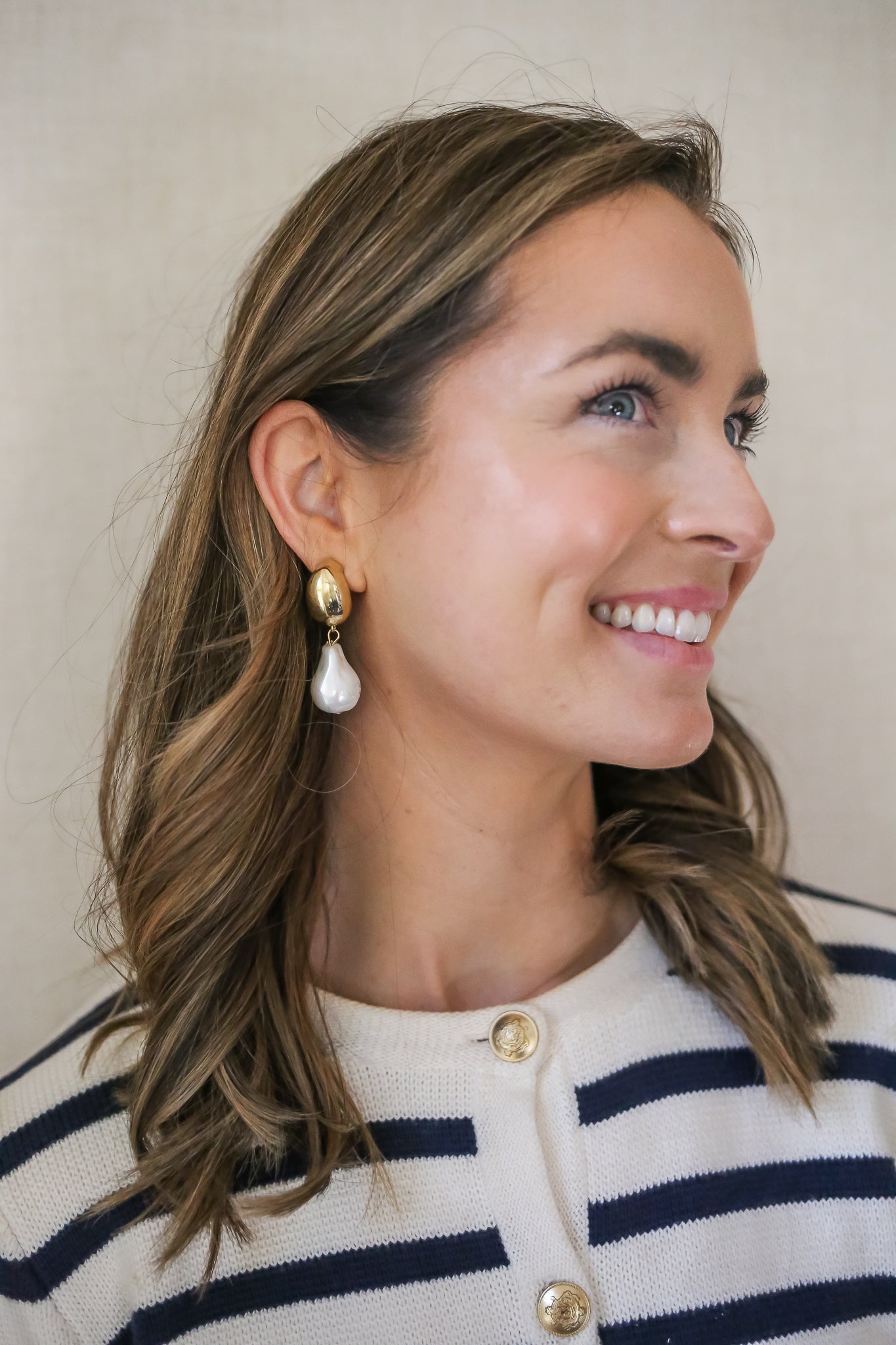 Woman with wavy brown hair wearing bold gold and pearl drop earrings, smiling while dressed in a navy and white striped cardigan with gold buttons, standing against a neutral beige background.