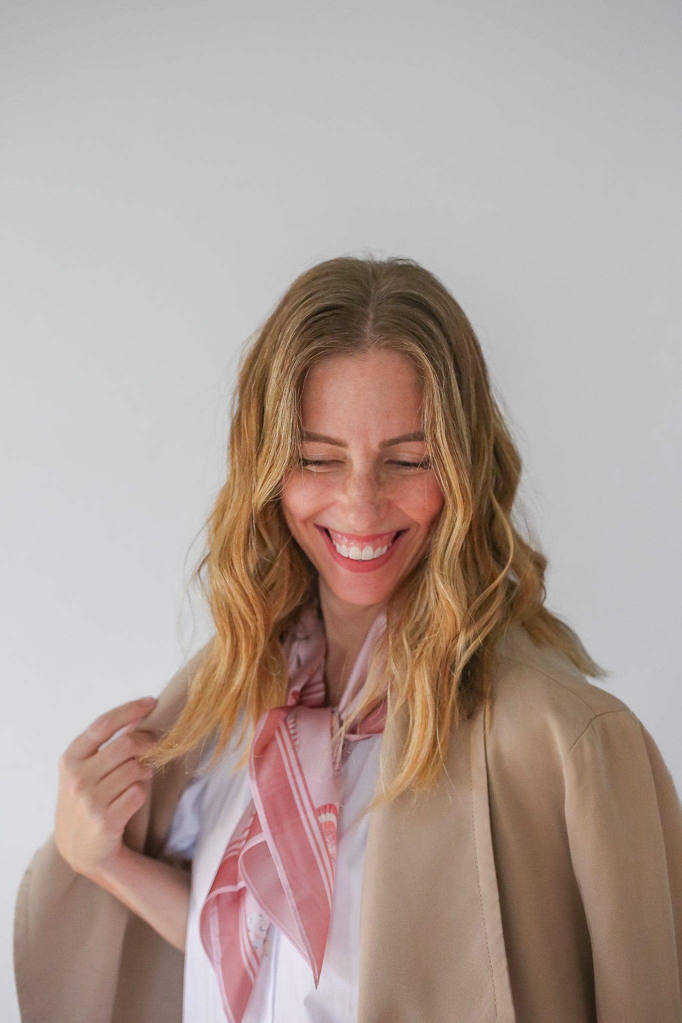 Woman laughing while holding the collar of a tan jacket paired with a pink silk scarf around her neck.