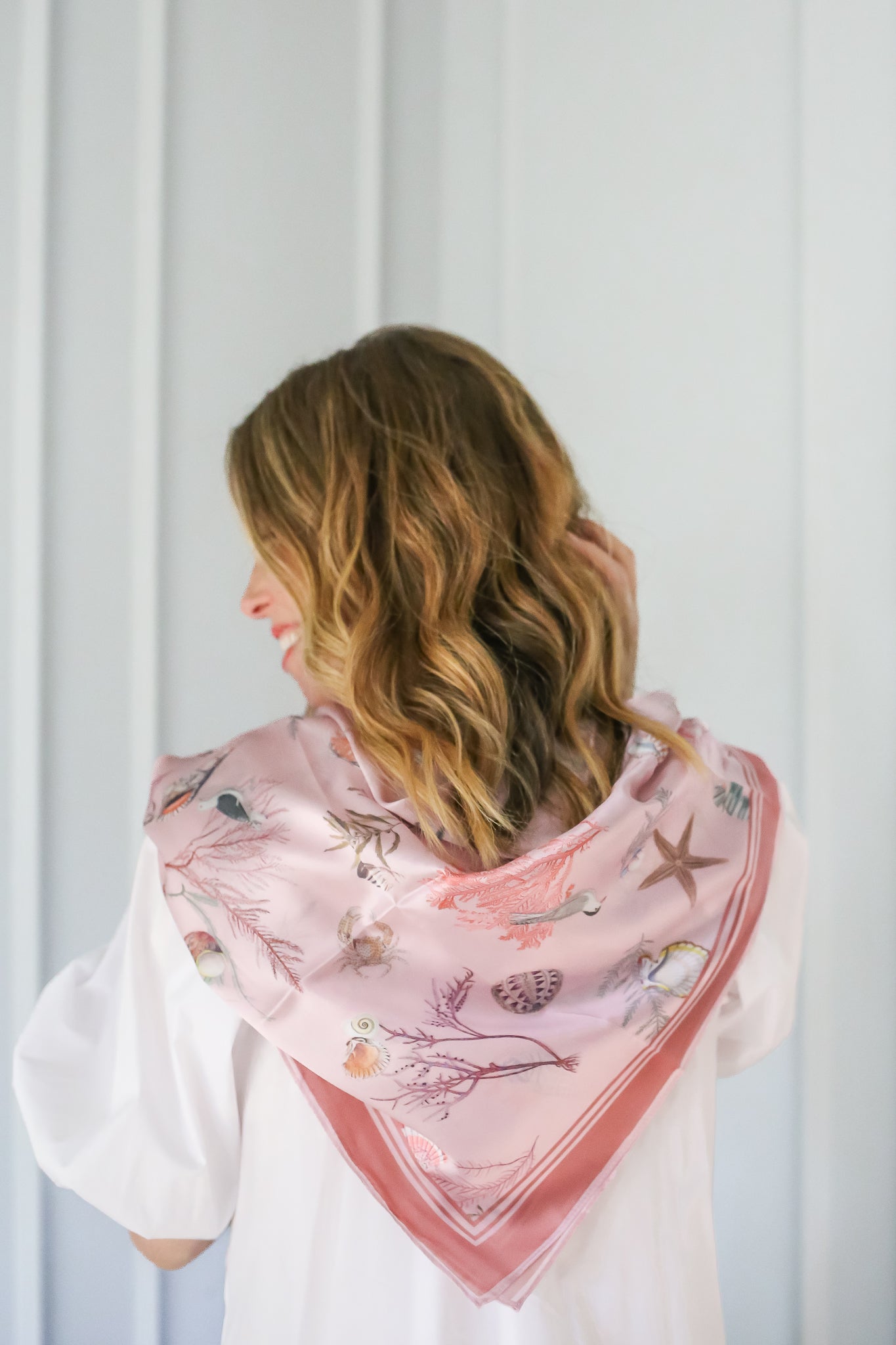 Back view of a woman with wavy blonde hair wearing a pink silk scarf adorned with seashell and starfish patterns.