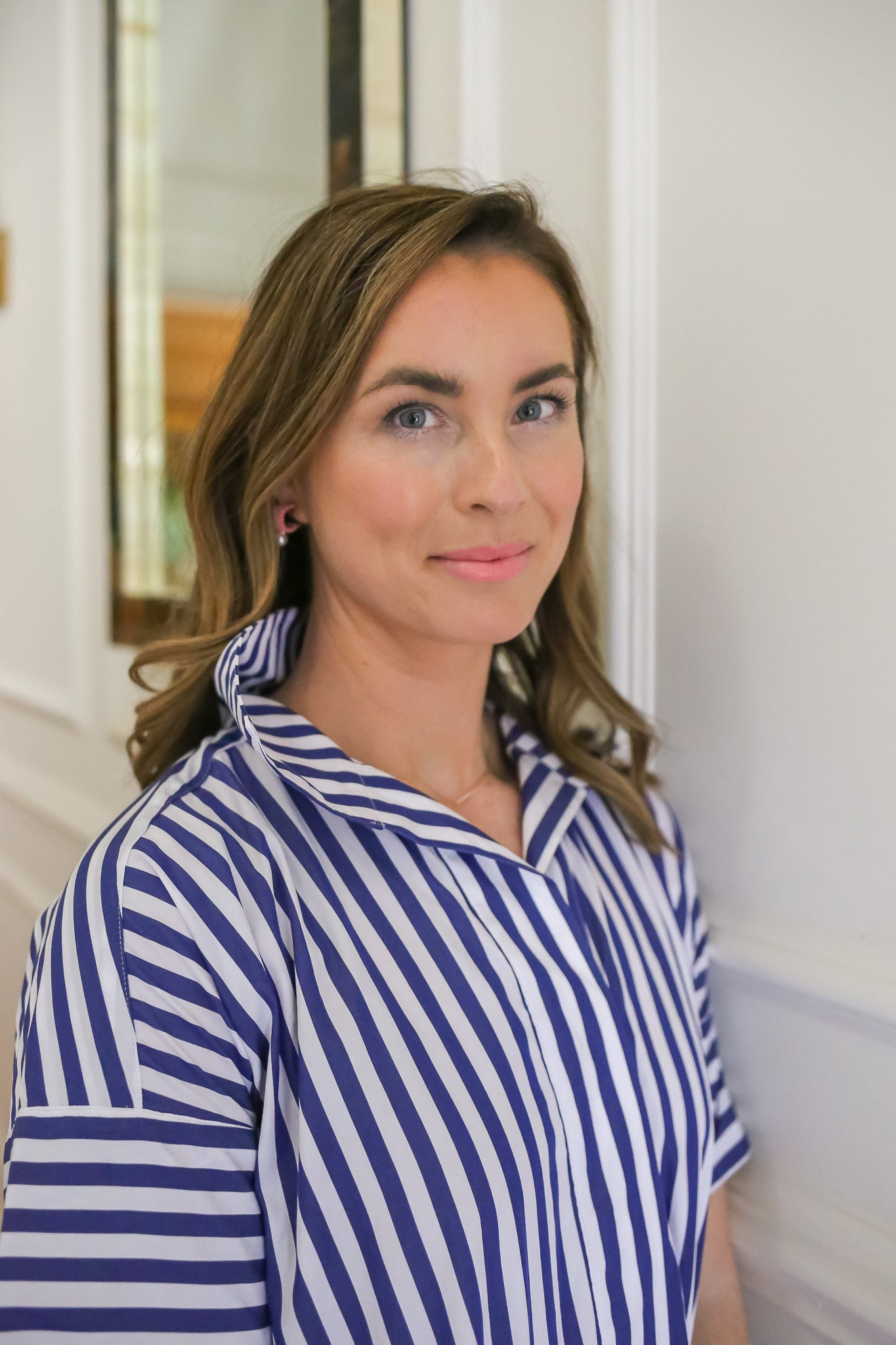 Portrait of Woman in Blue-and-White Striped Shirt: A confident woman with a neutral expression stands against a white wall, wearing a blue-and-white striped shirt and pink earrings, capturing effortless elegance and modern style.