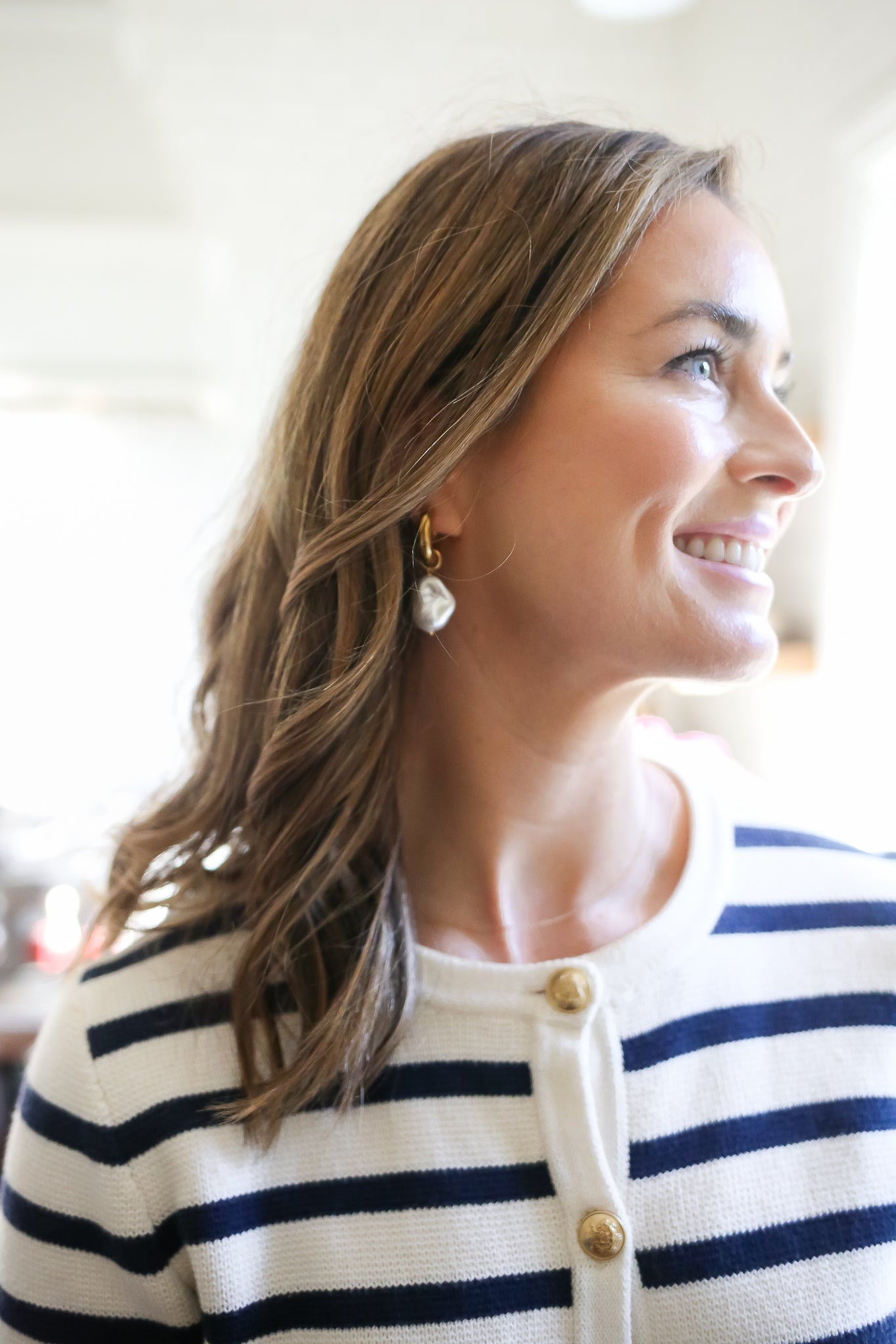 Side profile of a woman with wavy brown hair wearing gold statement earrings with a large pearl drop, paired with a navy and white striped cardigan, smiling as natural light illuminates her face.