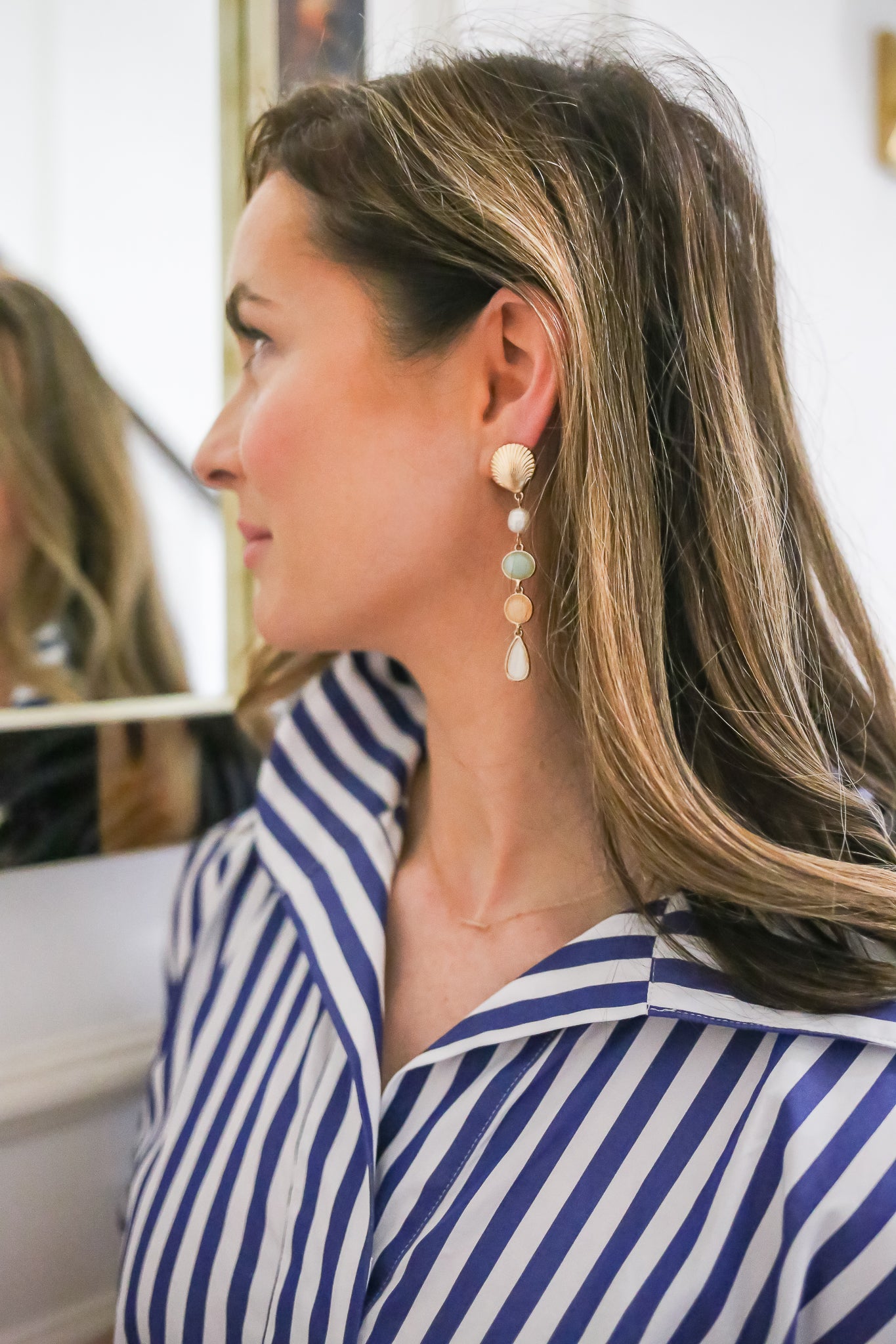 Side View of Woman Wearing Long Drop Earrings with Shell and Stone Accents: A woman with long, natural hair wears elegant drop earrings featuring a gold shell and pastel-colored stones, perfectly paired with a striped blue-and-white shirt.