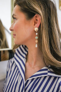 Side View of Woman Wearing Long Drop Earrings with Shell and Stone Accents: A woman with long, natural hair wears elegant drop earrings featuring a gold shell and pastel-colored stones, perfectly paired with a striped blue-and-white shirt.